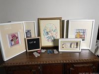 Five framed items displayed on wooden cabinet including four white frames and one gold frame with various artworks and small sculptures