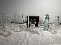 Photo showing five glass bottles, one clear pitcher, and two clear stemmed goblets arranged on a light surface with LOT 55 sign in background.