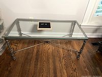 Rectangular glass top coffee table with metal tubular legs and crossbar supports, shown on hardwood floor, front angle.