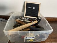 Clear plastic bin containing several used hammers and a variety of pliers and hand tools piled inside, showing wooden and rubber handles and metal parts. A sign labeled LOT 21 is placed behind the bin.