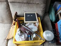 Yellow plastic crate on cushioned chair, filled with assorted nuts, bolts in bags and boxes, wooden-handled crowbar resting on top, round metallic light/reflector on the right side.