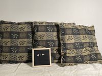 Three decorative pillows with creamy taupe and dark grey patterned covers in a square design, placed side by side with a sign indicating lot 44.