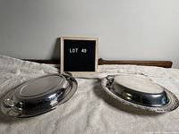 Top-down view of two oval silverplate chafing dishes with lids, one with smooth rim, the other with ornate border, placed on white fabric with wooden headboard background.