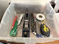 Clear plastic container holding assorted hand tools including tile cutter, utility knives, scissors, hand planer, marker, chalk line reel, tape, pencil, small level, and measuring tape.