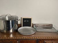 Wide view of all three kitchen cookware items placed on wooden surface: large stainless steel pot with glass lid, round tin pie plate with fluted edges, and rectangular glass lasagna pan.