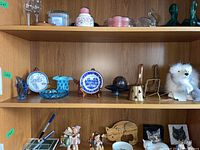 Shelf showing a mix of art glass items including fish figurines, a fish plate with stand, a glass ball, brass candle holder, white polar bear figurine, and other decorative glass pieces.