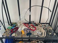 A wide shot of the costume jewelry lot showing various necklaces, earrings, and a brooch spread on a glass shelf with a black metal frame. Multiple colors and styles displayed, including faux pearl strands, gold-tone chains, and colorful resin pendants.