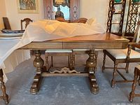 Front view of the wood dining table with the white padded cover partially removed, showing turned pedestal legs and carved base feet.