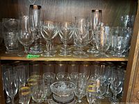 Two shelves holding various etched glassware items including wine glasses, coffee mugs, snifters, ashtrays, bowls and trays, all made of clear glass with etched decorative patterns.