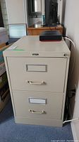 Front view of beige two drawer file cabinet with handles, label holders, and lock on top drawer; key seen on lock.