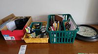 Photo showing multiple office supplies arranged in baskets and bins on a wooden surface, including scissors, pens, staplers, a calculator, and two clocks.