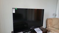 Front view of Samsung 46" LCD TV standing on wooden table with a beige chair in the background.