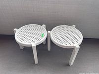 Two white plastic side tables with round perforated tops and three square legs, shown on a gray floor.