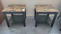 Pair of square matching side tables with laminate frames and faux marble tops on carpeted floor beside wall outlet.