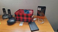 Photo shows two cordless phones with charging bases, Samsung Galaxy Tab 4 tablet with black case, red and black plaid storage box containing some accessories, Amazon Echo Dot smart speaker, and white charging cables on wood surface.