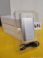 Three silver Allway Ewarmer handwarmers in clear plastic packaging with visible USB charging cable, photographed on a yellow surface with a stack of white boxes behind.