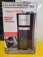 Front of Black+Decker coffee maker box showing single serve model, in black and silver, with 16 oz travel mug included. Handwritten note on box indicating missing filter basket and presence of a replacement basket to hold paper filters.