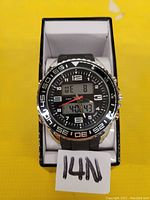 Front view of black watch with analog and digital display on a yellow surface in its box.