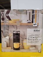 Photo of the Keurig K-Mini coffee maker in its original packaging showing the front of the box with an image of the product and brand labeling.