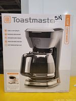 Front packaging photo of Toastmaster coffee maker showing product features including 12 cup capacity, removable brew basket, integrated cord storage, pause and serve function, and keep-warm heating plate.
