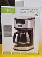 Front packaging view of TRU Programmable 12 Cup Coffee Maker showing brand, product image, and key features like digital control interface, 1-4 cup brew settings, and programmable brew timer.