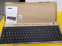 Photo showing Arteck wireless keyboard inside open box along with user manual. Keyboard is black with full key layout including numeric keypad. Box label indicates charging cable and USB receiver included inside.