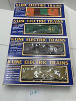 Four K-Line electric train boxcars in original packaging with window showing each boxcar's color and branding - orange/green, green, silver-gray, khaki. Boxes labeled with K-Line Electric Trains and O-27 gauge details.
