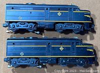 Side view of two Lionel Diesel model locomotives showing blue paint with different Erie logos, yellow striping, and visible wheels and couplers.