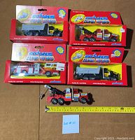 Five Majorette Super Movers diecast trucks, three boxed and two loose, various types including dump trucks, fire truck, and tow truck. Measuring tape shows approximate size of loose tow truck.