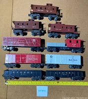 Nine mixed Lionel model train cars displayed on a flat surface with a measuring tape showing approximate length. Various types and colors including cabooses, boxcars, and gondolas.