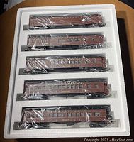 Five passenger train cars in Tuscan brown and black packed in styrofoam tray, each wrapped in protective plastic.
