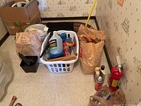 Full view showing black floor mats, laundry basket with blue fluid and spray cans, mop, brown bags with supplies, and fire extinguisher.