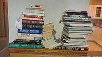 Two stacks of over 40 hardcover and coffee table books on a wooden table, titles visible on spine include 'Armand Hammer', 'Iacocca', 'Leaving Home', 'The Builders', and 'Great Cars of the 20th Century'.