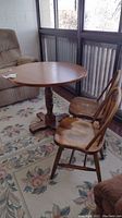 Full view of the round wooden dining table with two matching wooden chairs placed on a floral rug by windows.