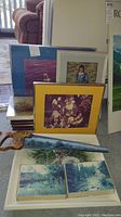 Assortment of framed photos and prints stacked together with a painted hand saw laid across them, showing various mat colors and themes.