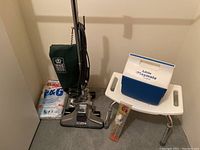 Photo of a Royal upright vacuum cleaner with green fabric bag cover, pack of vacuum bags, Little Playmate cooler on a white shower bench with adjustable legs, and Hoppe's cleaning kit on the bench.