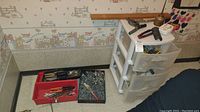 Overview of the lot showing various hand tools in red and black trays on the floor next to a white plastic three drawer storage unit with additional tools and hardware on top.