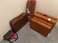 View of two brown vintage Samsonite hard side suitcases, vintage sleeping bag in brown carrying bag, and four soft side vinyl luggage pieces in brown and red.