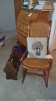 Walnut wooden rocking chair with cane seat and backrest, floral pillow on seat, and wooden magazine rack placed on floor next to chair.