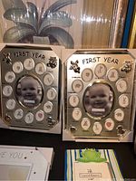 Two silver-tone metal baby first year milestone photo frames with labeled monthly slots and a central photo of a baby.