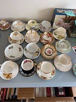 Photo showing multiple teacup sets with matching saucers arranged on table, displaying a wide variety of floral and gold-trimmed patterns.