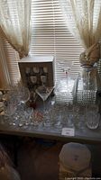 Photo of various glassware and ceramic tea set on table including wine glasses, martini glasses, shot glasses, and decorative glass containers