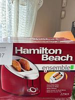 Photo of the unopened Hamilton Beach toaster box showing the product image and branding on a red and white background, with bagels in the toaster slots.