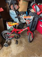 Full view of red boys' bike with training wheels, flame decals, and training wheels attached, placed indoors on concrete floor.