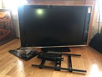 Sony Bravia 52-inch flat screen TV model KDL-52XBR3 shown with disassembled black metal wall mount bracket and hardware in plastic bags on floor in front of TV.