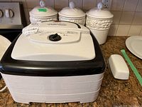 Photo of Nesco American Harvest food dehydrator with dial control, placed on kitchen counter with three white ceramic covered canisters and butter dish behind it.