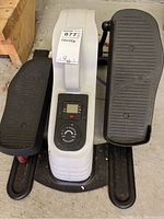 Front view of mini stepper showing two large black foot pedals, central white control unit with digital display and control knob.