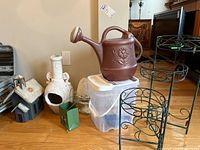 Garden decor items including neuxtral colored vases, brown plastic watering can, black metal plant stand with three shelves, and foldable metal bags on wooden floor