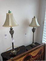 View of the pair of matching metal table lamps with fabric shades on a patterned surface against a white wall.
