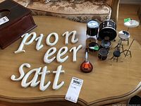 Miniature drum set with black Zildjian drums, cymbals, small mandolin figure, and white wall art letters on a wooden table.